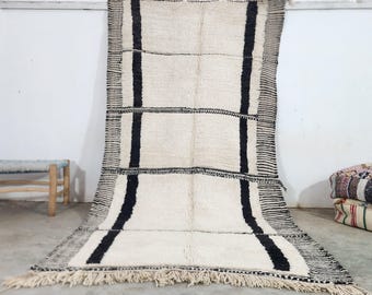 May include: A white and black Moroccan rug with a geometric pattern. The rug has a fringe border and is made of wool.