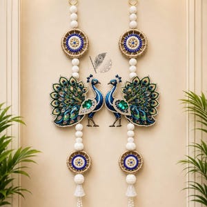 Op de afbeelding: Een paar verticale wandhangers met twee blauwe pauwen met uitgespreide staarten, geaccentueerd door groene en blauwe juwelen. Elk stuk bevat witte kralenstrengen, cirkelvormige geweven frames en witte kwastjes aan de onderkant.