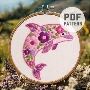 May include: Embroidered dolphin design in a wooden hoop. The dolphin is filled with pink, purple, and green floral embroidery. The text "PDF PATTERN" is in a white circle in the upper right corner. The background is blurred.