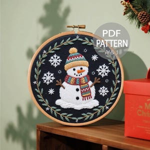 May include: Embroidered snowman in a wooden hoop. The snowman wears a colorful hat and scarf, with snowflake accents on a dark blue background. The text "PDF PATTERN A&J" is visible.