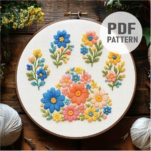 May include: Embroidered floral paw print design in a wooden embroidery hoop. The design features blue, orange, yellow, and pink flowers with green leaves. The text "PDF PATTERN" is in a white circle.