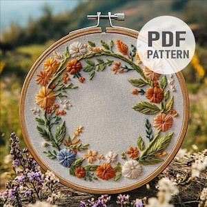 Puede incluir: Un aro de bordado con una corona de flores naranja, amarilla, blanca y azul sobre un fondo de tela beige. El aro es de madera y tiene un acabado natural. El bordado está hecho en un estilo delicado e intrincado. La imagen incluye el texto "PDF PATTERN".