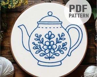 Puede incluir: Un bastidor de bordado con tela blanca, con un diseño de tetera bordada en azul con detalles florales. El texto "PDF PATTERN" está en un círculo blanco. El bastidor está sobre un fondo de madera.