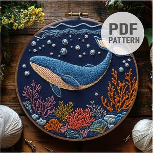 May include: Embroidered hoop art featuring a blue whale swimming through a coral reef with bubbles. The whale is stitched with blue and pink thread, and the coral is stitched with orange, pink, and green thread. The background is a dark blue fabric.