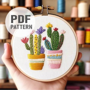 K&ouml;nnte beinhalten: Ein PDF-Muster zum Sticken mit zwei Topfpflanzen, die Kakteen mit bunten Bl&uuml;ten darstellen. Die Kakteen sind gr&uuml;n mit gelben und rosa Akzenten, und die T&ouml;pfe sind gelb und rosa mit bunten Streifen.