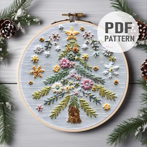 Puede incluir: Diseño de árbol de Navidad bordado en un aro de madera. El árbol está decorado con flores y copos de nieve de colores. El texto "PDF PATTERN" está en un círculo blanco. El fondo incluye agujas de pino y una piña.