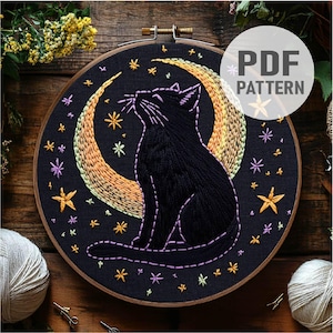 Puede incluir: Arte de bordado en aro con un gato negro sentado bajo una luna creciente con un degradado arcoíris. El fondo es una tela azul oscuro con estrellas amarillas, verdes y moradas.