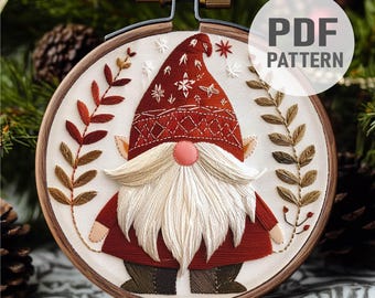 May include: Embroidered Christmas gnome pattern in a wooden hoop. The gnome is wearing a red hat with white snowflakes and a red and white patterned sweater. The gnome has a white beard and a red nose. The pattern is surrounded by green leaves.