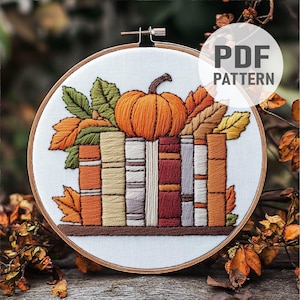 May include: Embroidered hoop art featuring a stack of books with a pumpkin on top, surrounded by autumn leaves. The hoop is made of wood and has a white fabric background.