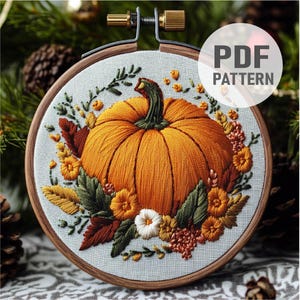 Może przedstawiać: Haftowany obraz w obręczy przedstawiający szczegółowy dynię ze łodygą i liśćmi, otoczony kompozycją kwiatową z białych, żółtych i pomarańczowych kwiatów. Obręcz jest drewniana i ma jasnoniebieski materiałowy podkład.