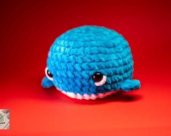 May include: A handmade, plush blue whale toy with white accents. The whale has large, black eyes with white highlights and small fins. The toy is set against a vibrant red background, creating a striking contrast.