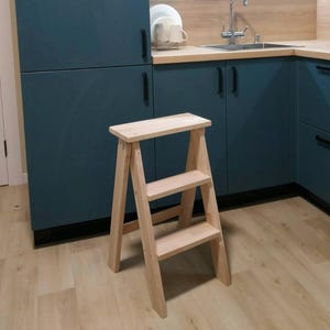 Puede incluir: Un pequeño taburete de madera sin terminar con tres escalones. El taburete está hecho de madera de color claro y tiene un diseño simple y funcional. Es adecuado para alcanzar estantes o armarios más altos en una cocina u otra área.
