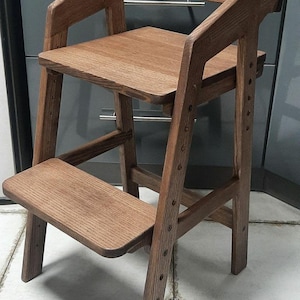 Puede incluir: Silla alta de madera con acabado natural. La silla tiene asiento, reposapiés y reposabrazos. La altura del asiento y el reposapiés es ajustable. La silla es de madera y tiene un diseño clásico.