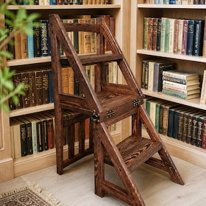 May include: A dark stained wooden library step stool with a folding design. The step stool has two steps and a seat, with brass hinges. The background features a bookshelf filled with books.