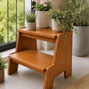 Könnte beinhalten: Ein zweistufiger Tritthocker aus Holz mit warmbrauner Oberfläche. Auf der oberen Stufe stehen drei Topfpflanzen: ein kleiner grauer Topf mit Lavendel, ein großer weißer gerippter Topf mit rosa Blüten und ein heller Topf mit Efeu.