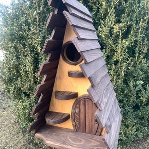 Puede incluir: Una caprichosa pajarera de madera con un tejado de tejas marrón oscuro y un cuerpo marrón claro. Cuenta con un agujero de entrada redondo, pequeños escalones de madera y una puerta arqueada en miniatura. La pajarera está montada en un poste de madera.