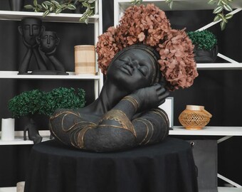 Op de afbeelding: Zwarte buste sculptuur van een vrouw met gesloten ogen, versierd met een krans van gedroogde bloemen. De sculptuur heeft gouden accenten op de armen en staat op een zwarte tafel. Decoratieve items staan op witte planken op de achtergrond.