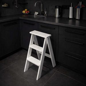 Puede incluir: Un taburete plegable blanco de tres escalones. El taburete de madera tiene un diseño sencillo. Se muestra en una cocina con armarios oscuros y una encimera negra. El taburete mide aproximadamente 61 cm de altura.