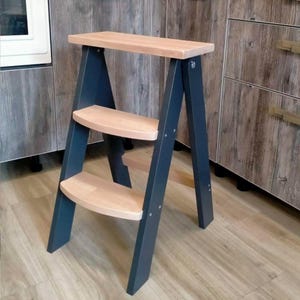 Puede incluir: Un taburete de madera con una parte superior y escalones de color claro. El marco está pintado de un gris azulado oscuro. El taburete tiene tres escalones y está diseñado para alcanzar lugares más altos.