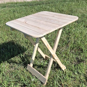 Puede incluir: Pequeña mesa plegable de madera clara, con tapa rectangular y patas cruzadas. La mesa está sobre un campo de hierba. Sus dimensiones son de aproximadamente 61 cm de largo, 46 cm de ancho y 51 cm de alto.