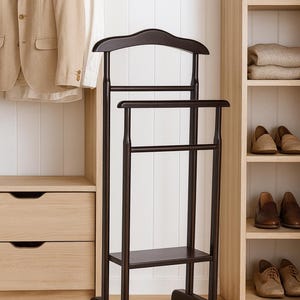 Op de afbeelding: Een donkerbruine houten kledingstandaard met een gebogen hanger voor jasjes, een horizontale stang voor broeken en een onderste plank voor accessoires. De vrijstaande structuur wordt ondersteund door twee gebogen voeten voor het georganiseerd ophangen van kleding.