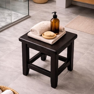 May include: A dark brown wooden stool in a bathroom setting. The stool holds a stack of beige towels, a wooden brush, and a brown glass bottle with a black pump. A woven basket with white towels is in the foreground.