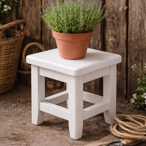 May include: A white wooden plant stand with a square top supports a terracotta pot filled with lavender. The stand is on a dirt surface, with a basket and gardening tools in the background. The wooden background adds a rustic touch.