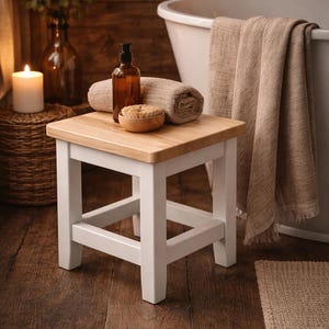 Könnte beinhalten: Ein kleiner Holzschemel mit weißem Gestell und einer Tischplatte aus Naturholz. Ein gerolltes Handtuch, eine Flasche und eine Holzschale liegen obenauf. Die Szene spielt in einem Badezimmer mit Badewanne und Kerzen.