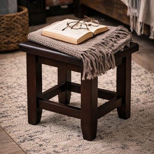 May include: A dark brown wooden stool with a square top. A book, glasses, and a fringed throw blanket rest on top. The stool sits on a patterned rug, suggesting a cozy reading nook.