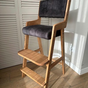 Könnte beinhalten: Ein hölzerner Hochstuhl mit dunkelgrau gepolstertem Sitz und Rückenlehne. Der Stuhl hat eine Fußstütze und Armlehnen. Das Holz hat eine natürliche Oberfläche. Der Stuhl ist für ein Kind konzipiert.