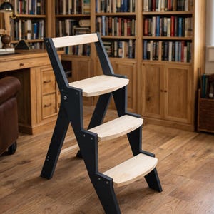 Op de afbeelding: Een houten opstapje met drie treden. Het frame is zwart en de treden zijn van lichtgekleurd hout. Het opstapje staat in een kamer met boekenkasten en een parketvloer.