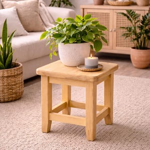 May include: A small, light-colored wooden stool with a square top. A potted plant with green leaves sits on top, next to a gray candle on a small, round coaster. The stool is in a living room setting with other plants and furniture.