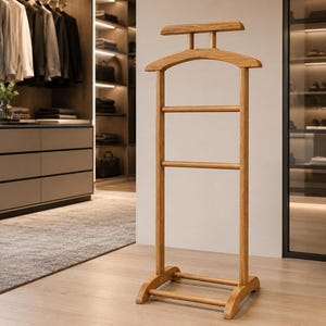 Könnte beinhalten: Ein freistehender Herrendiener aus hellem Holz mit einem breiten, geformten Kleiderbügel für Jacken, einer horizontalen Stange für Hosen und einer zweiten unteren Stange, alles getragen von einem stabilen Fuß mit zwei geschwungenen Beinen.