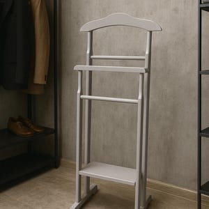 May include: A light gray wooden valet stand with a curved top, a shelf, and two horizontal bars for hanging clothes. The stand is designed to hold garments and accessories, and is placed in a room with a neutral color scheme.