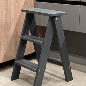 May include: A grey wooden step stool with three steps. The stool has a folding design with a wide top step and two lower steps. The legs are angled for stability. The stool is painted a solid grey colour and is suitable for reaching high places.