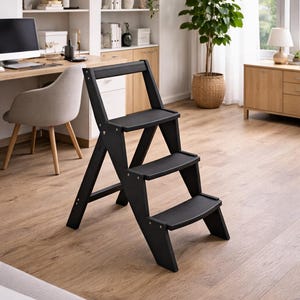 Puede incluir: Un taburete plegable negro con tres escalones. Fabricado en madera, está diseñado para uso en interiores. La imagen muestra el taburete sobre un suelo de madera clara. Se aprecia un escritorio, una silla y estanterías en el fondo.