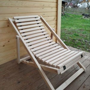 Puede incluir: Una tumbona plegable de madera clara con asiento y respaldo de listones. La silla está hecha de madera sin terminar y está diseñada para uso en exteriores. La silla está colocada en una terraza de madera.