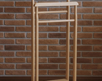 May include: A light-colored wooden valet stand with a hanger, a horizontal bar, and a small shelf. The stand is made of light-colored wood and stands on a wooden floor against a brick wall.