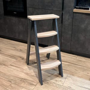 Puede incluir: Un taburete de madera con estructura gris y tres escalones de madera natural. El escalón superior es rectangular y los inferiores son curvos. El taburete está diseñado para alcanzar estantes o armarios más altos.