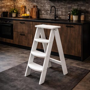 Puede incluir: Un taburete de madera blanco con tres escalones y una plataforma superior. El taburete está colocado sobre una alfombra gris oscura en un entorno de cocina. El taburete mide aproximadamente 76 cm de altura.