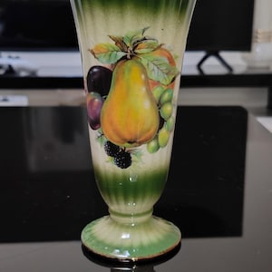 May include: A vintage ceramic vase with a flared top and pedestal base. The vase has a green and cream colour scheme with a painted design of fruits, including a pear, grapes, and plums. The rim has a gold trim.