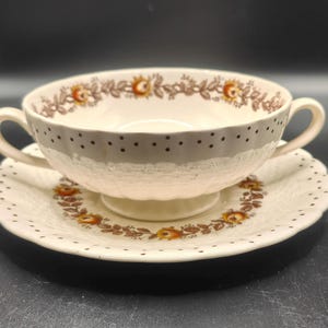 May include: Cream-colored soup bowl with two handles, sitting on a matching saucer. The bowl and saucer feature a floral and dot pattern in brown and orange. The bowl has a textured surface.