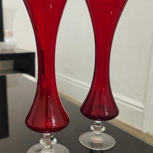 May include: Two tall, ruby red glass vases with flared tops and a bulbous midsection. The vases sit on clear, circular bases. The elegant design and rich colour make them suitable for decorative purposes.