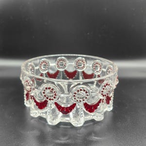 May include: A clear glass bowl with a decorative red pattern around the rim. The bowl has a circular design with red accents and a scalloped edge. The bowl is likely for decorative purposes.