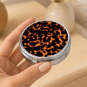 May include: A round, silver-toned metal compact mirror featuring a vibrant orange and black leopard print pattern on the lid. The compact has a polished metallic finish around the circular edge and a secure hinge mechanism for opening and closing.