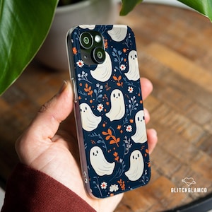 May include: A navy blue phone case featuring a pattern of white cartoon ghosts, orange leaves, and white flowers. The case has a clear border and is held in a hand. The phone's camera lenses are visible.
