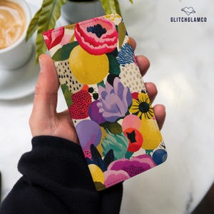 Könnte beinhalten: Eine rechteckige Handyhülle mit einem lebendigen, mehrfarbigen Blumen- und abstrakten Muster in Rosa, Gelb, Violett, Rot und Grün. Das Design zeigt verschiedene Blumenformen und gepunktete Texturen auf einem cremefarbenen Hintergrund mit GLITCHGLAMCO-Text.