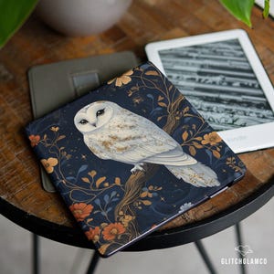 Op de afbeelding: Een Kindle-hoesje met een wit uil-ontwerp op een donkerblauwe achtergrond met bloemendetails. De hoes ligt op een houten tafel naast een Kindle e-reader. Het ontwerp bevat oranje en gele bloemen.