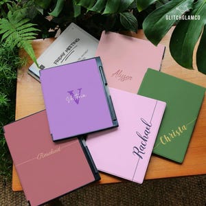 Può includere: Una collezione di quaderni personalizzati in vari colori, tra cui rosa, viola e verde, con nomi come "Alyssa" e "Victoria" stampati sulle copertine. Un quaderno mostra il testo "Friday Meeting".
