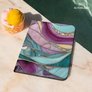 Pu&ograve; includere: Una custodia per tablet con un colorato design a vetrata. La custodia presenta sfumature di viola, verde acqua e oro, con una finitura lucida. Il design &egrave; astratto, con linee e forme curve. La custodia &egrave; su una superficie di marmo.